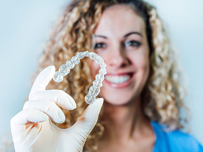 The image shows a person holding up a transparent orthodontic retainer with visible teeth impressions against a white background, smiling at the camera.