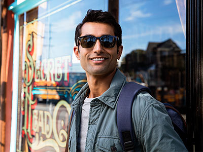 A man wearing sunglasses and a backpack stands outside a storefront, with a sign visible behind him.